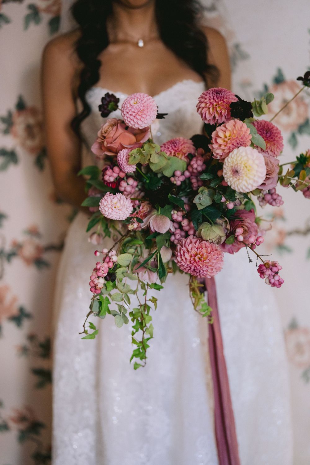 Bride in a strapless white lace gown holds a lush pink dahlia bridal bouquet with greenery and a long mauve ribbon against floral wallpaper.