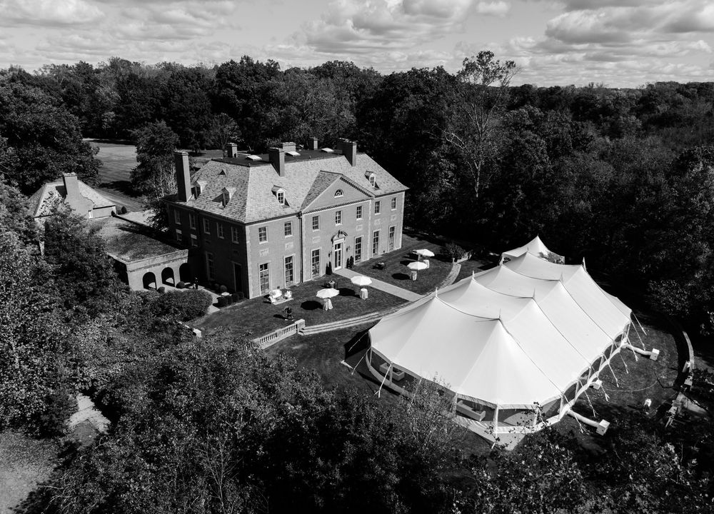 Aerial black and white view of a grand brick mansion surrounded by trees with a large white event tent set up on the lawn beside it.