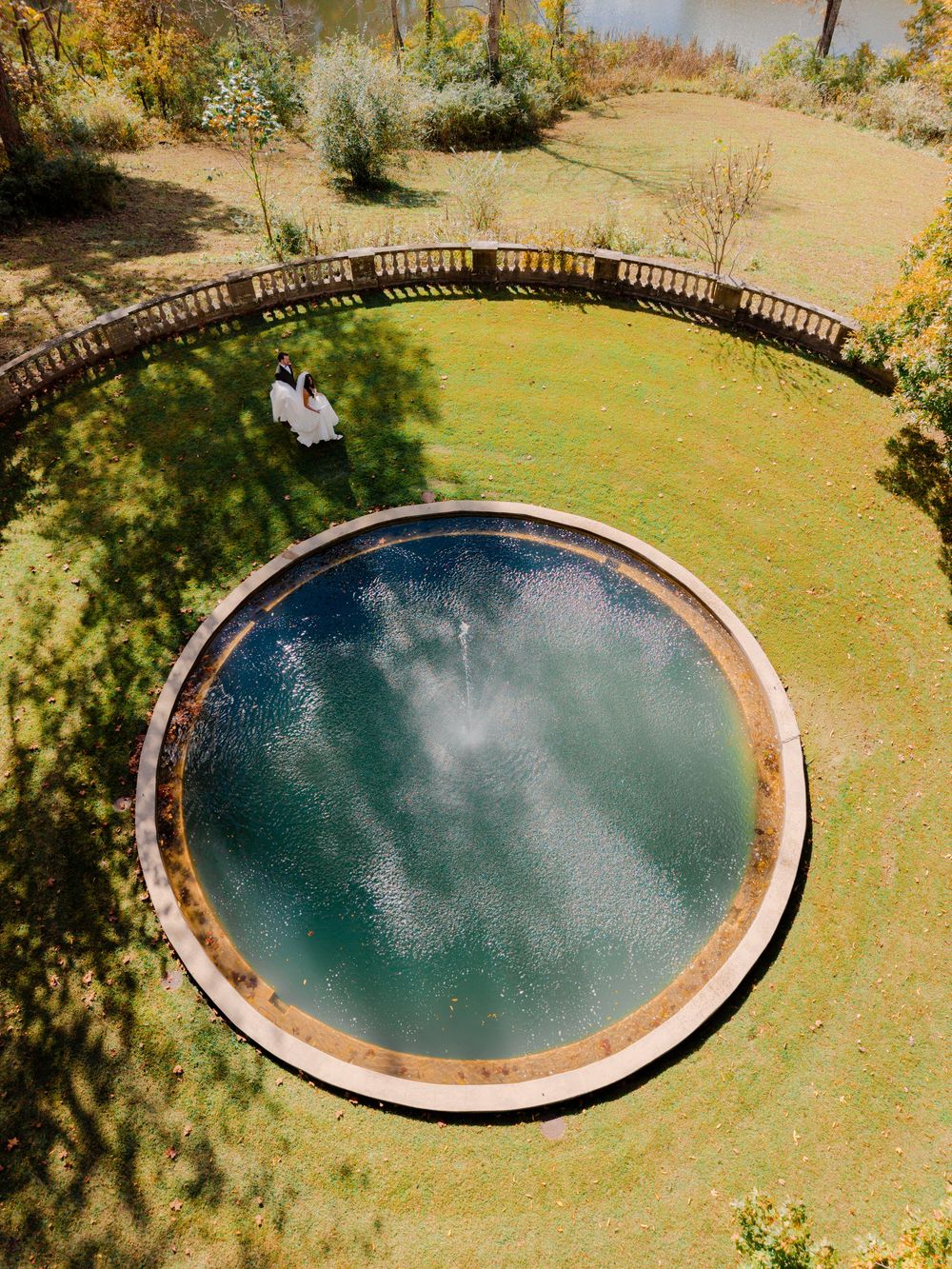 Aerial view of a round fountain pool with a bubbling center on a green lawn, with a couple in white standing by a curved stone balustrade.