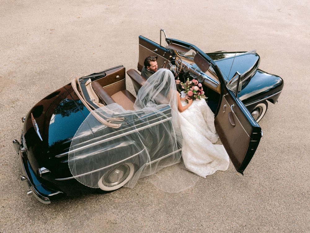 Bride in a white gown with a long veil sits in a vintage black convertible while the groom sits beside her, seen from above.