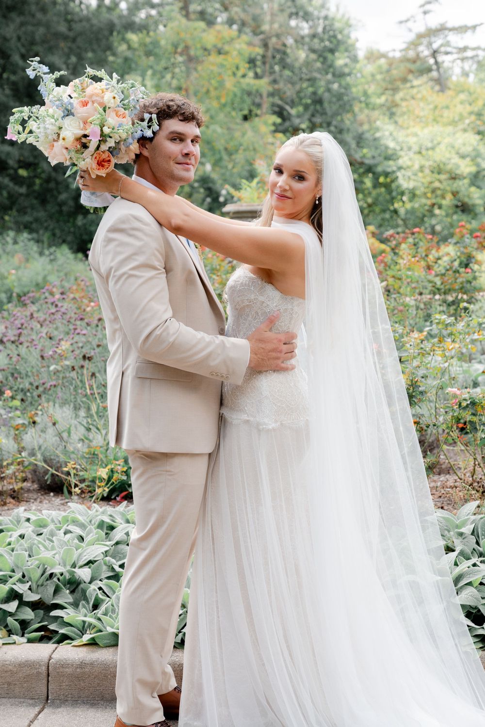 Bride in white gown and veil holds bouquet behind groom in tan suit, posing in lush garden setting.