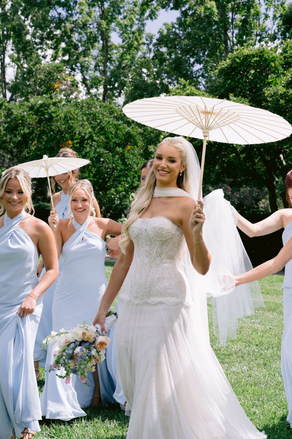 Bride in white lace gown holds parasol with bridesmaids in light blue dresses outdoors on sunny wedding day.