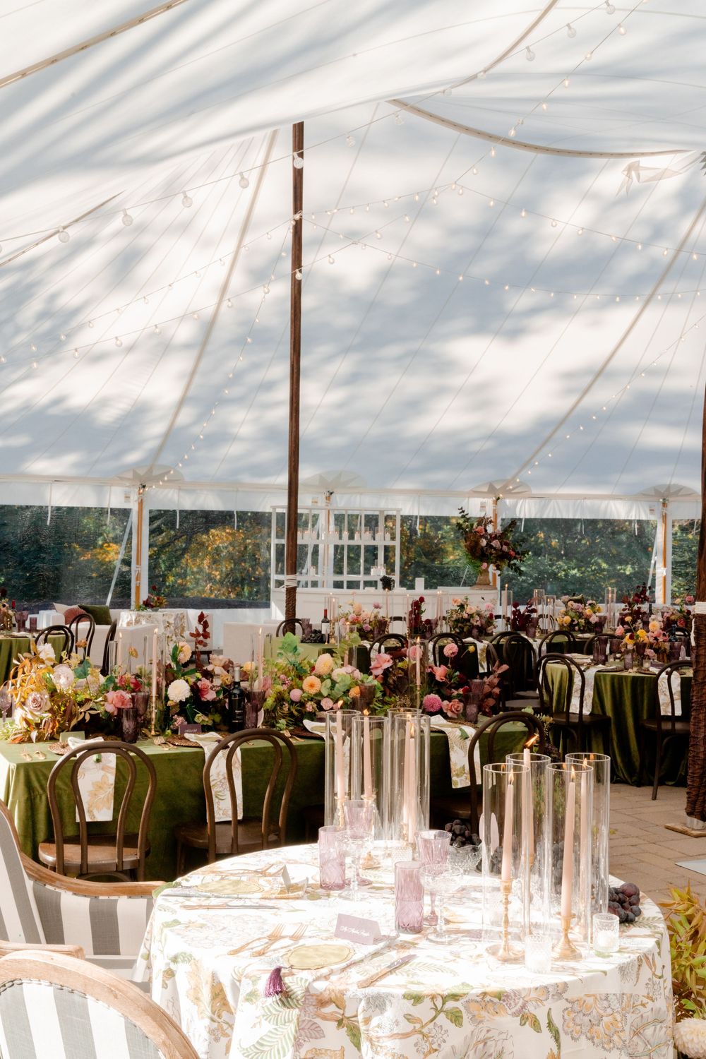 Elegant white tent wedding reception with floral centerpieces, taper candles, glassware, and green and floral linens on tables.