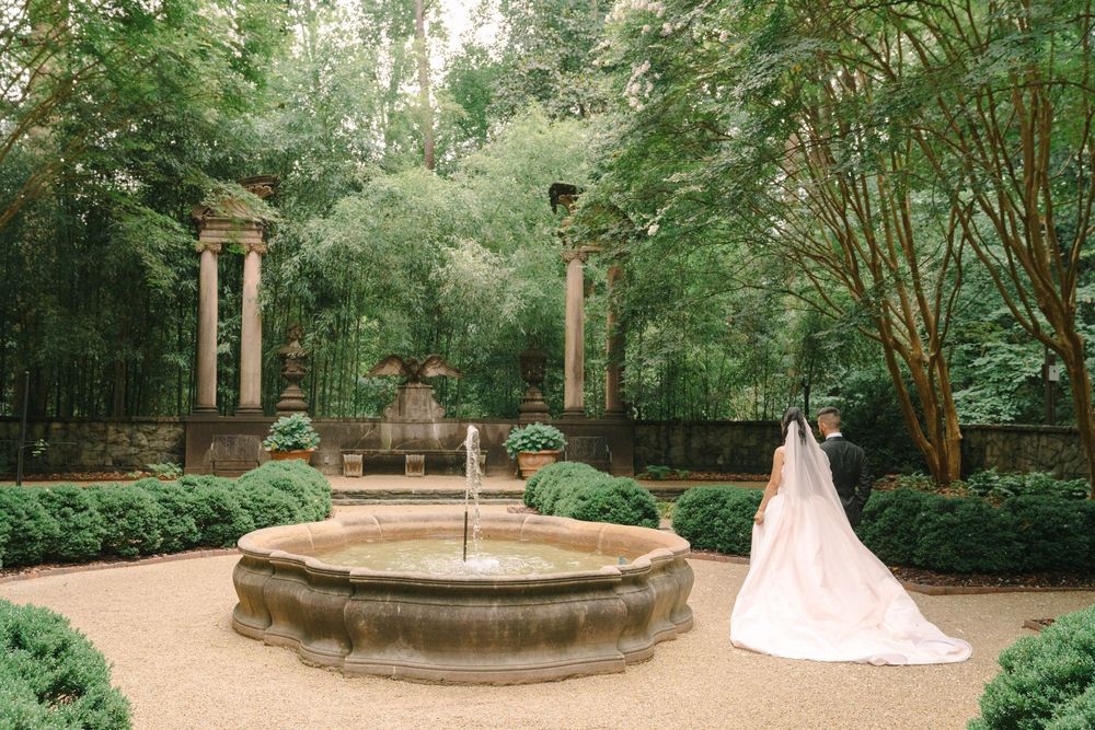Bride in flowing gown with cathedral veil stands by stone fountain in lush formal garden with columns.
