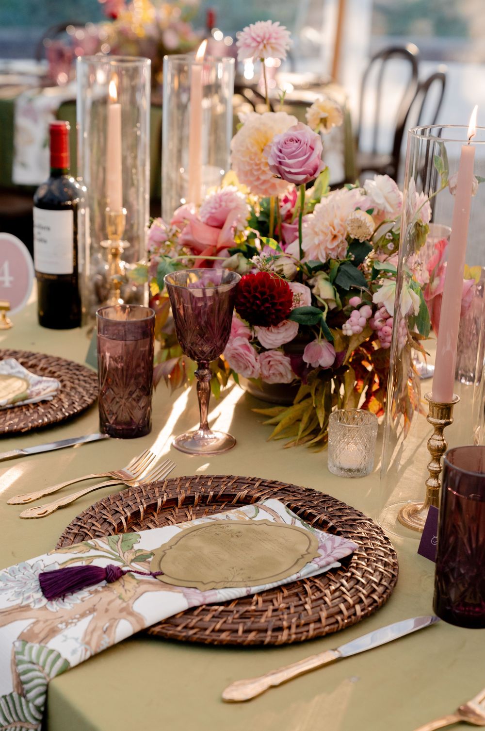 Elegant wedding table setting with a woven rattan charger, floral napkin with tassel, gold cutlery, taper candles and pink rose centerpiece.