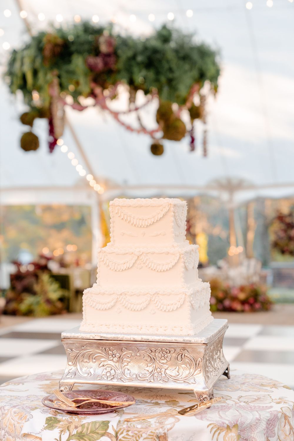 Three-tier square white wedding cake with piped swag details on an ornate silver stand beneath a hanging floral installation in a tent.