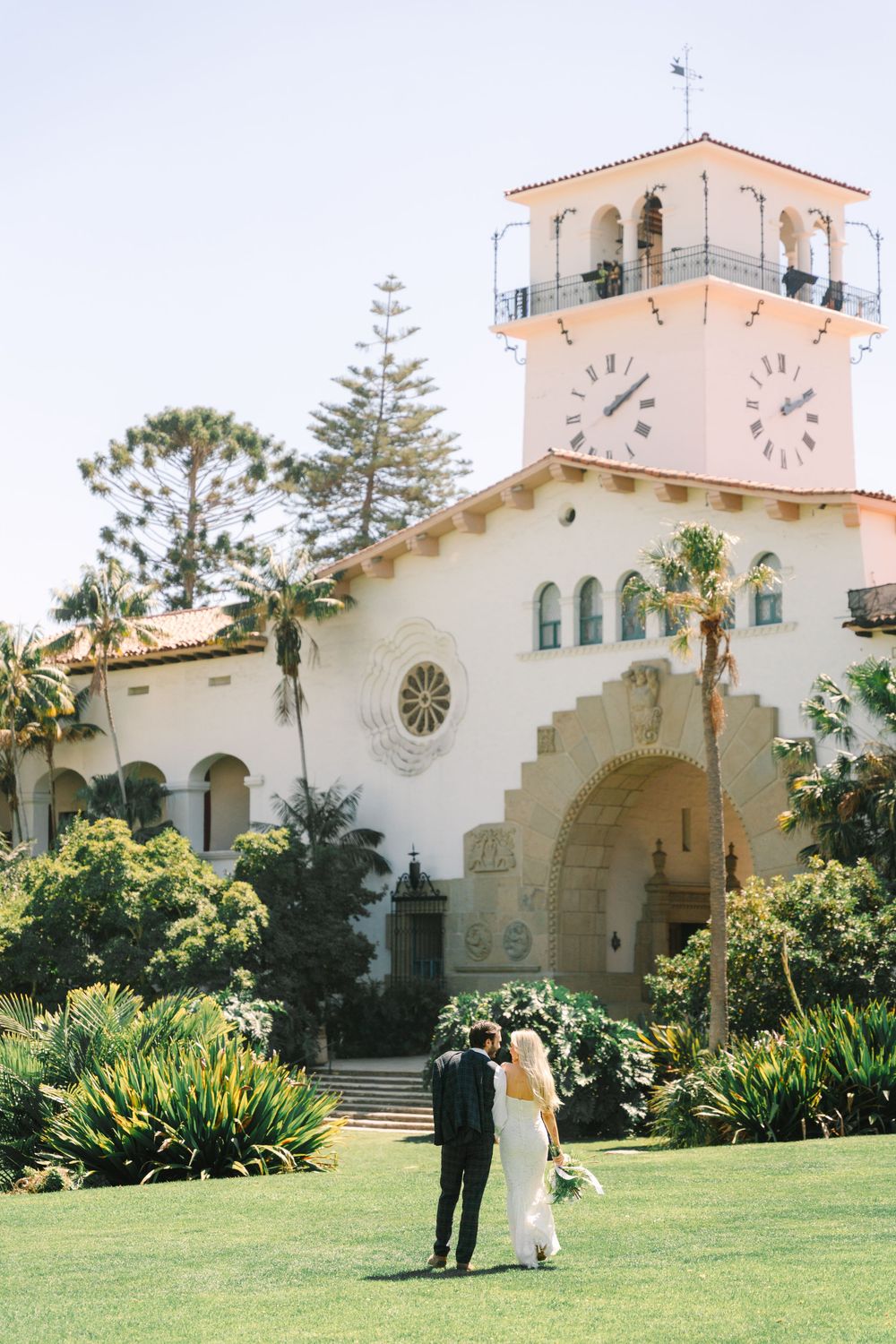 Bride and groom walking toward a white Spanish colonial building with clock tower surrounded by lush greenery.