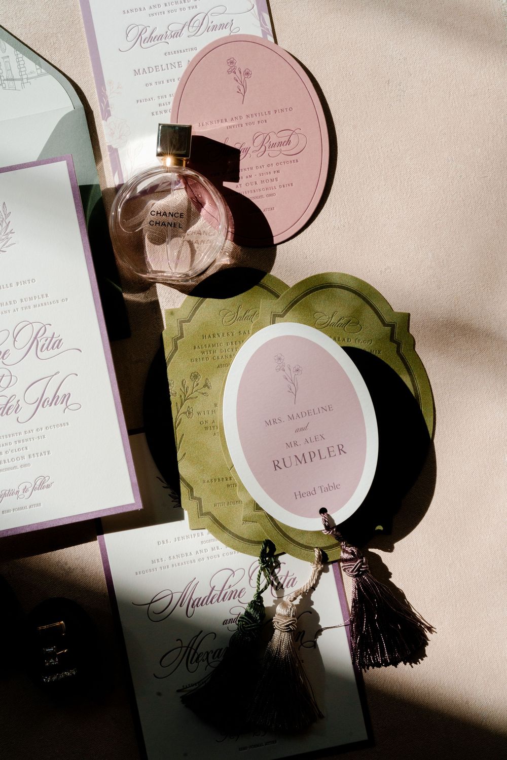 Elegant wedding stationery flat lay with blush and olive green oval place cards, tassels, rehearsal dinner invitation and Chanel perfume.