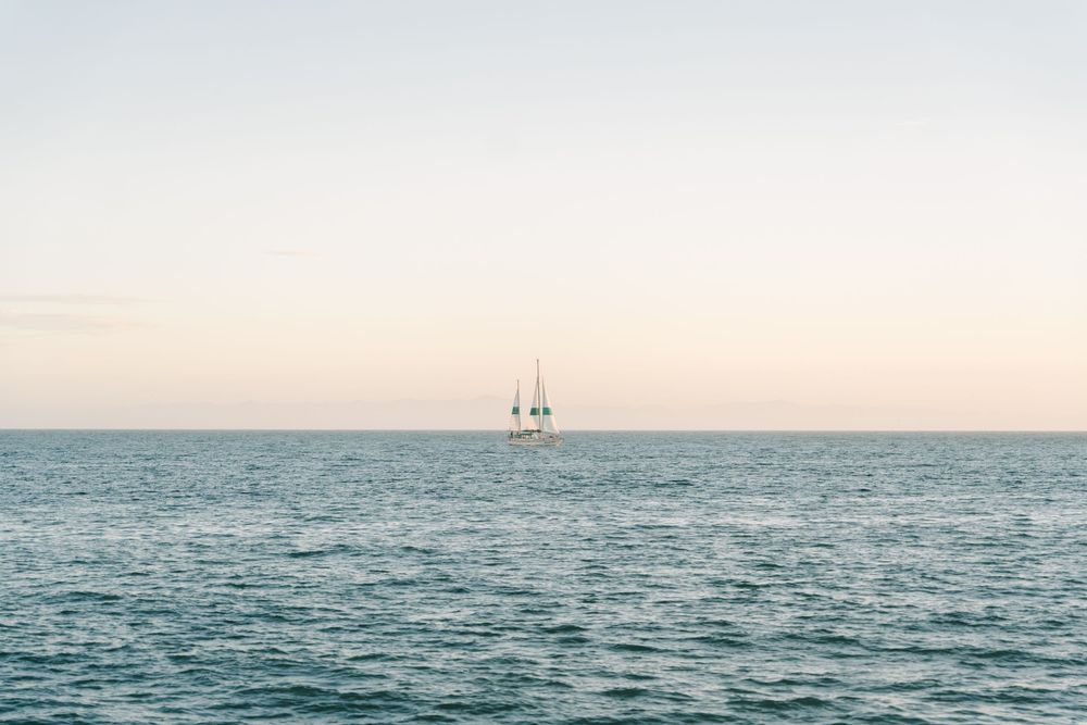 A lone sailboat glides across calm blue ocean waters under a soft pastel sky at dusk.