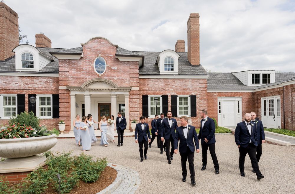 Wedding party walks outside elegant brick mansion venue, groomsmen in tuxedos and bridesmaids in white gowns on sunny day.