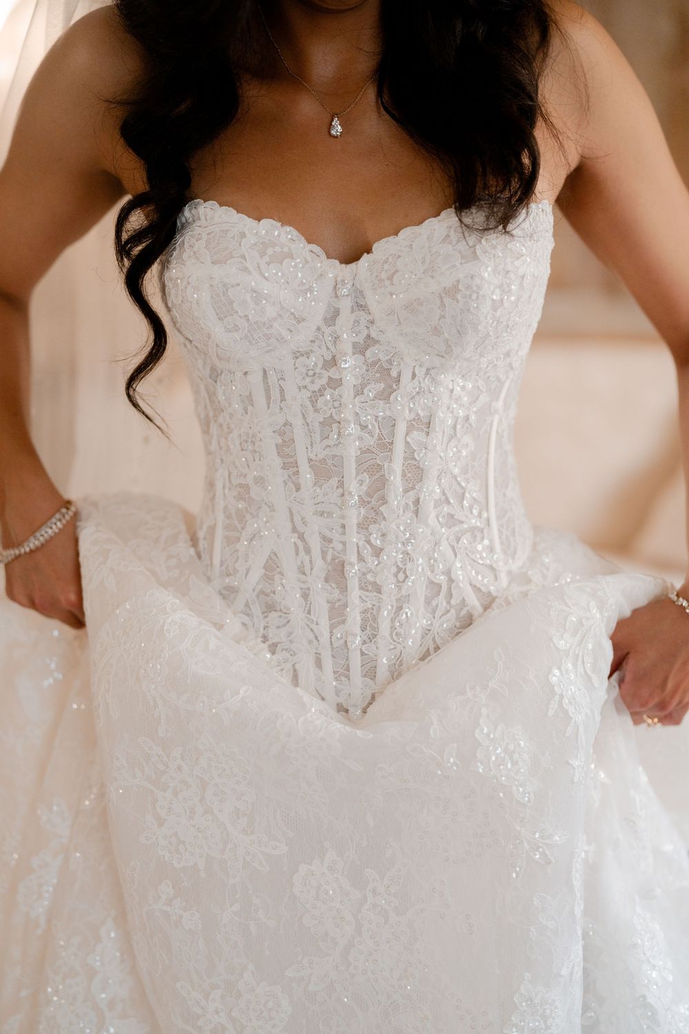 Bride in a strapless ivory lace corset wedding gown with sweetheart neckline and full tulle skirt, wearing a pendant necklace and bracelet.