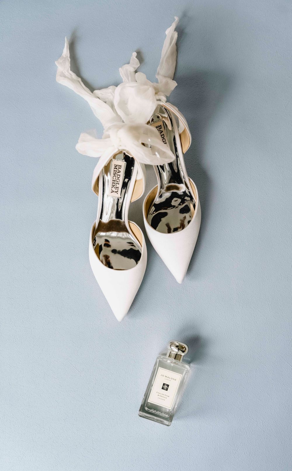 White Badgley Mischka bridal heels with floral ribbon ties beside a Jo Malone perfume bottle on a gray surface.