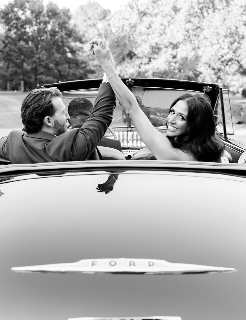 Black and white photo of a smiling couple raising joined hands in the back of a vintage Ford convertible, surrounded by trees.