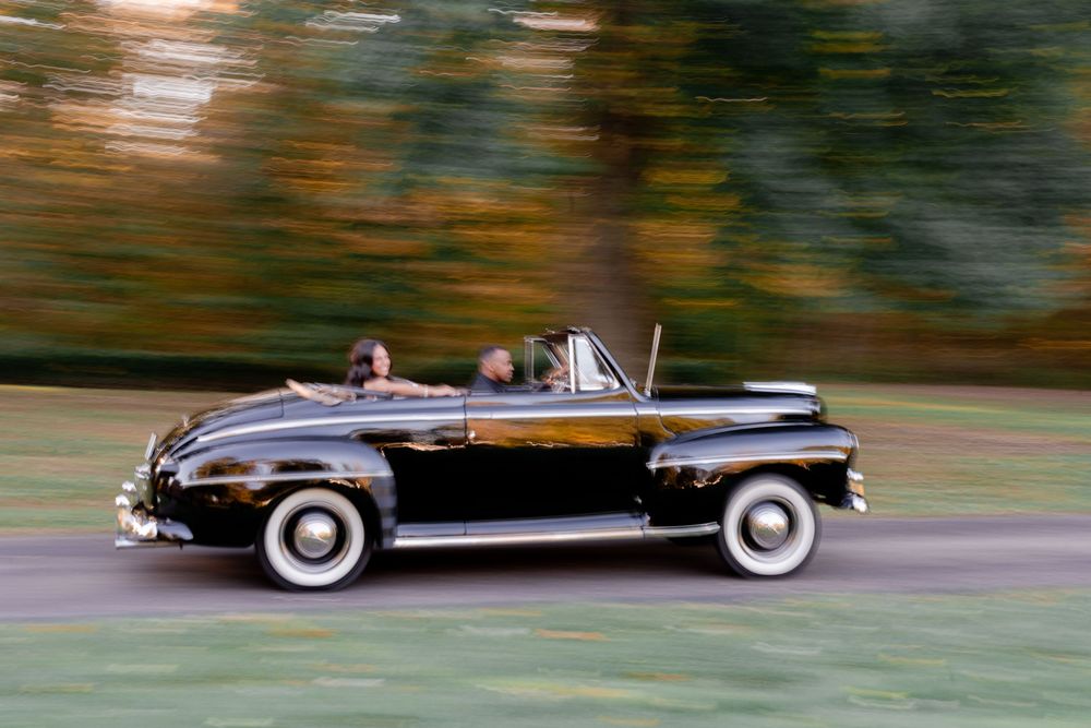 Two people ride in a black vintage convertible with white wall tires along a tree-lined road, with motion blur in the autumn background.