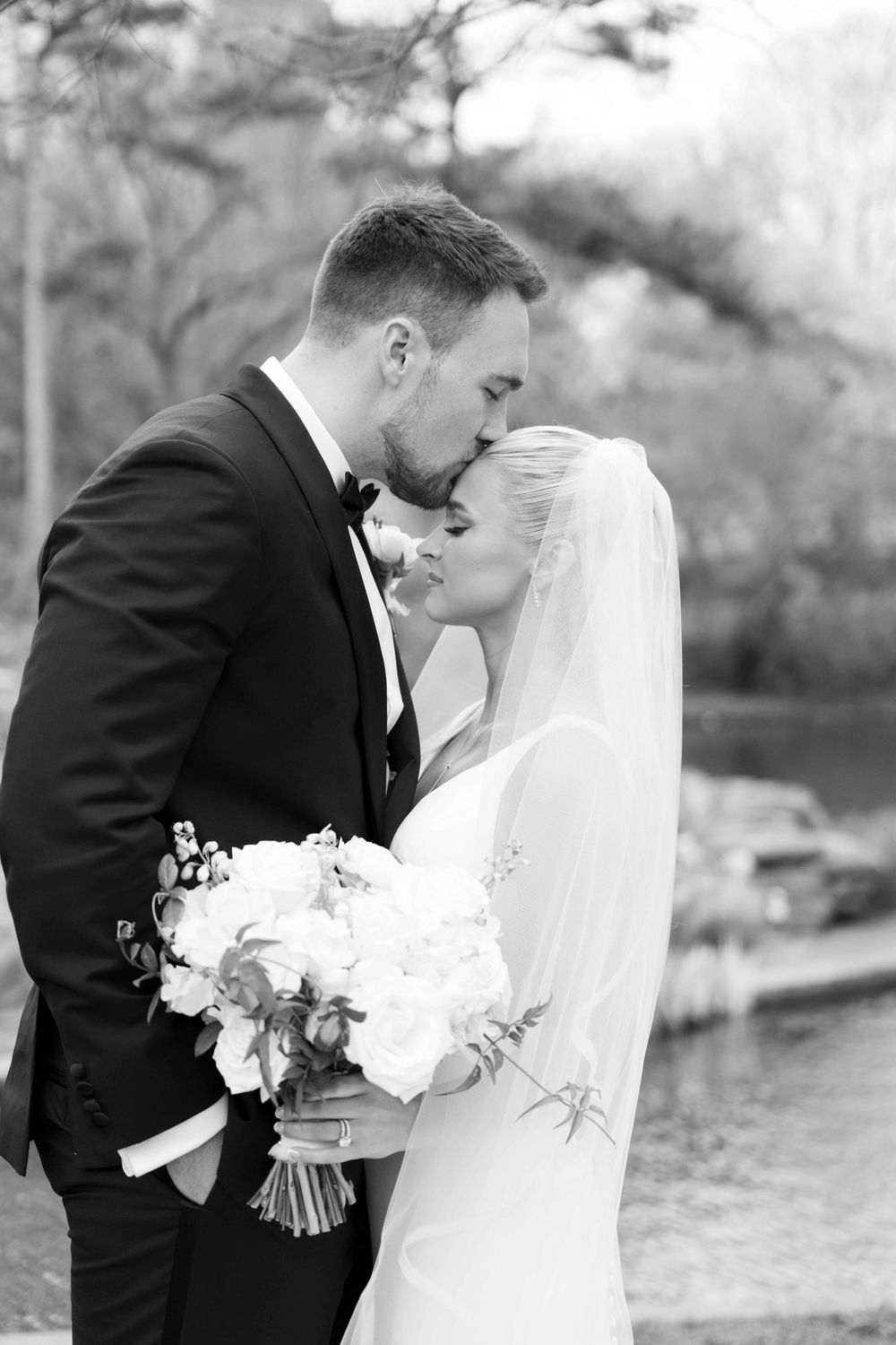 Groom in black tuxedo kissing bride in white gown and veil while she holds a white rose bouquet.