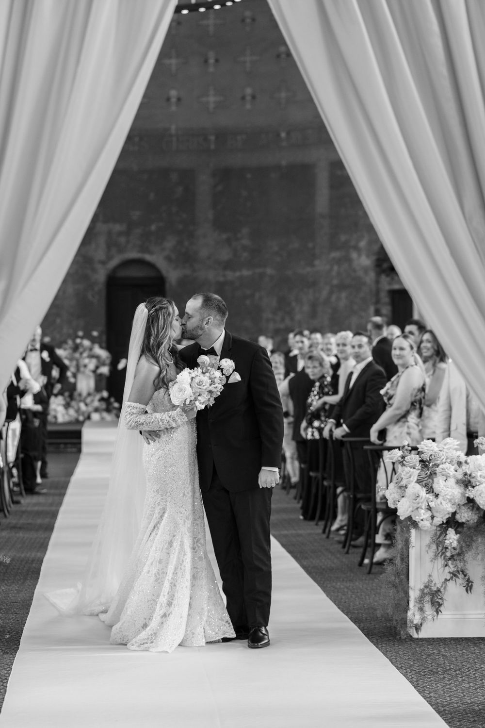 Black and white photo of bride and groom sharing a kiss at the altar during their elegant wedding ceremony.
