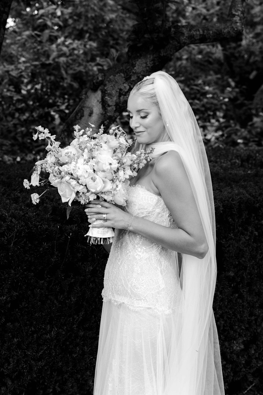 Black and white photo of a bride in lace gown and veil smelling her floral bouquet outdoors.