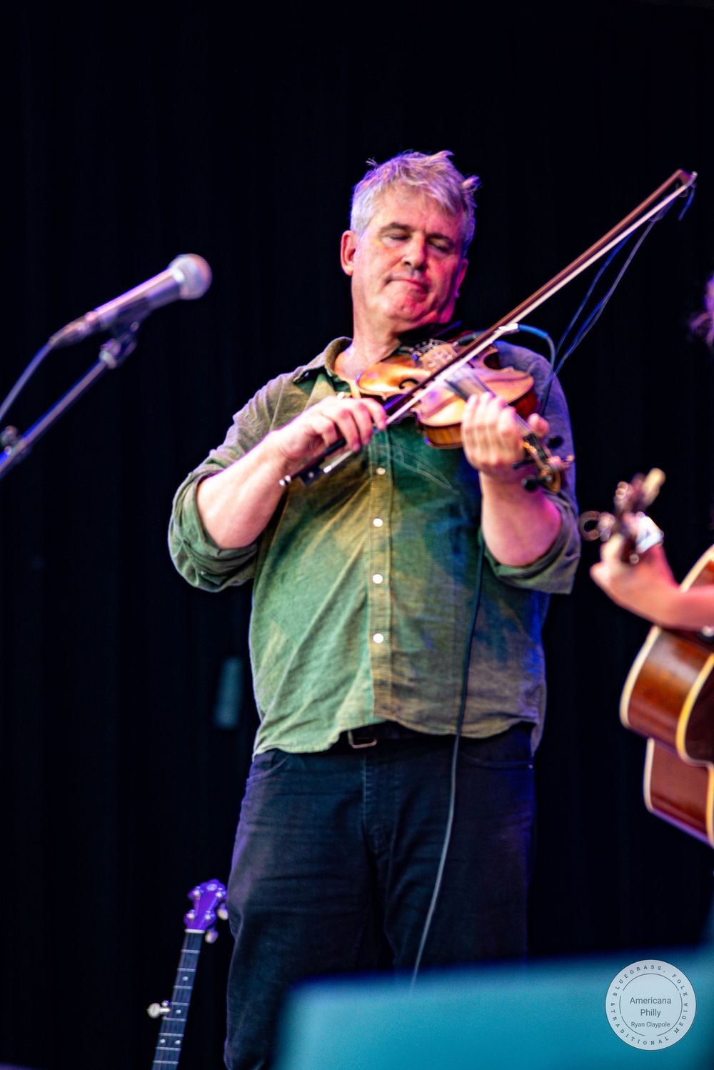 Middle-aged man playing violin on stage, wearing green shirt, performing live music concert with microphone and guitarist visible.