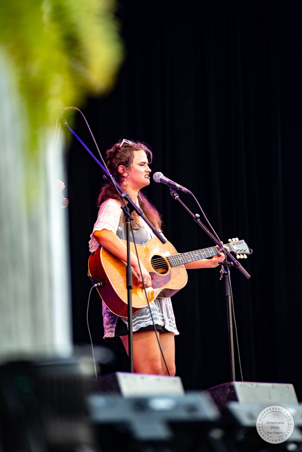 Young woman playing acoustic guitar and singing into microphone on outdoor stage during live music festival performance.