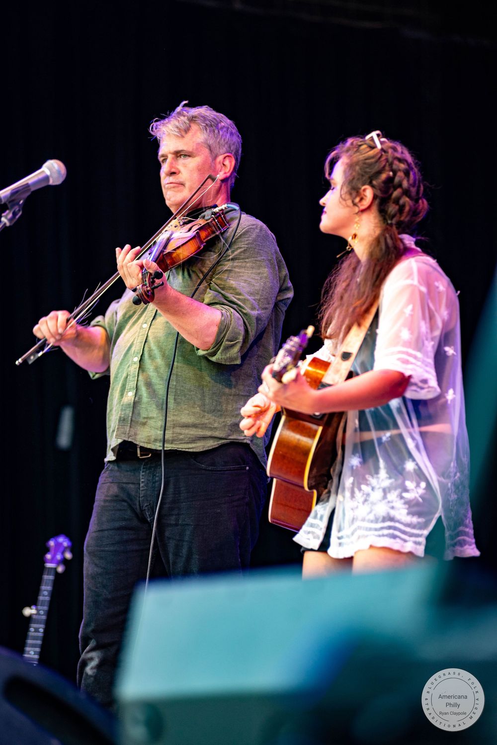 A man playing violin and a woman playing acoustic guitar perform together on an outdoor stage at a live music event.