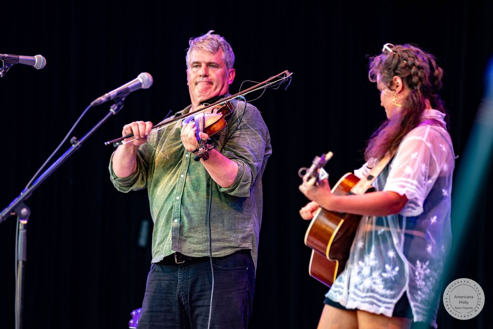 Two musicians perform on stage, a man playing violin and a woman strumming acoustic guitar under colorful stage lights.