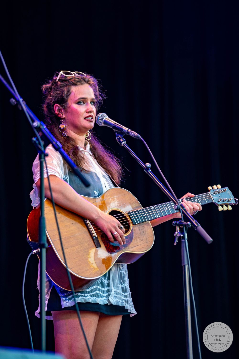 Young woman performing on stage playing acoustic guitar and singing into a microphone under purple stage lighting.