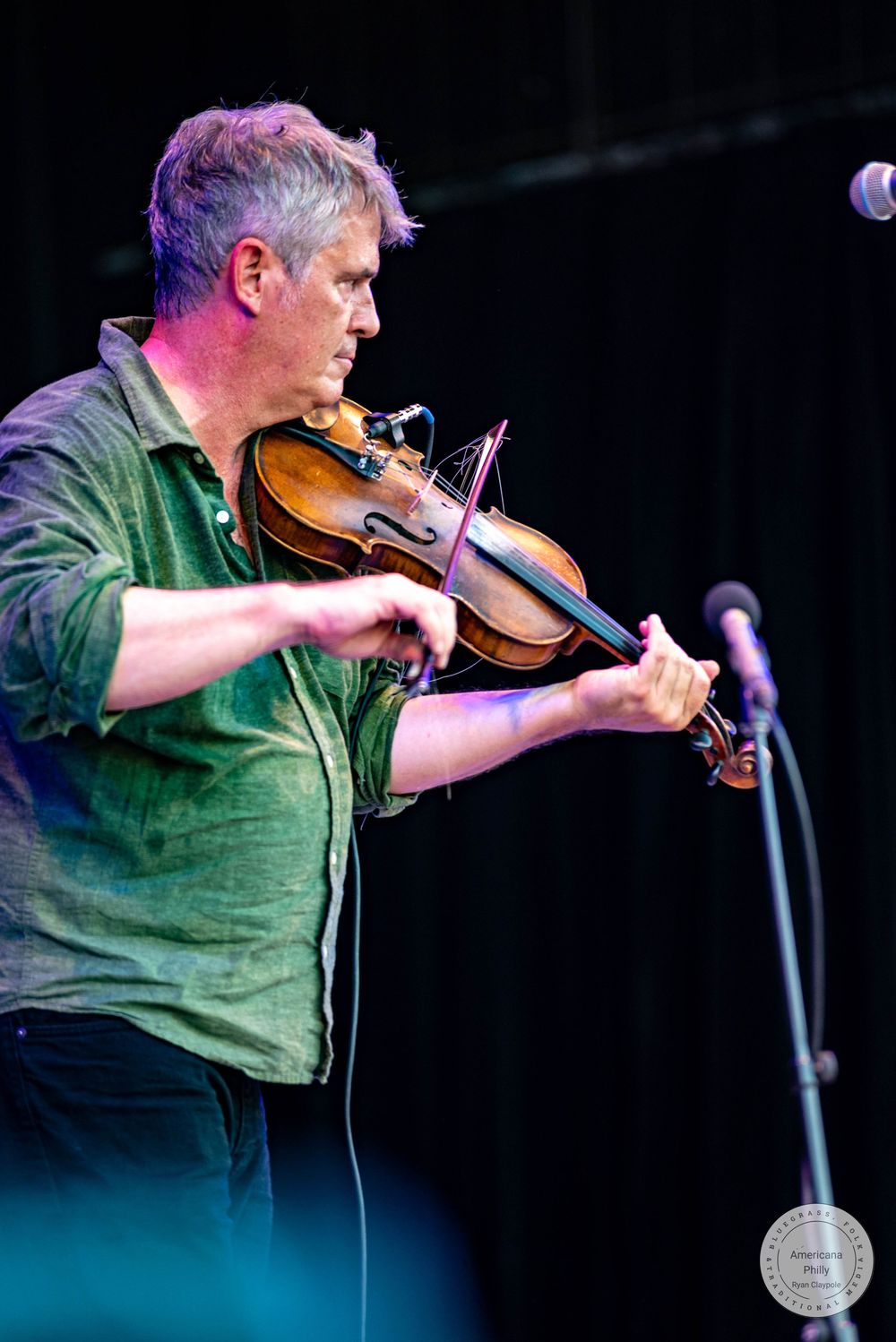 A man plays violin on stage, wearing a green shirt, performing live at an outdoor music festival.