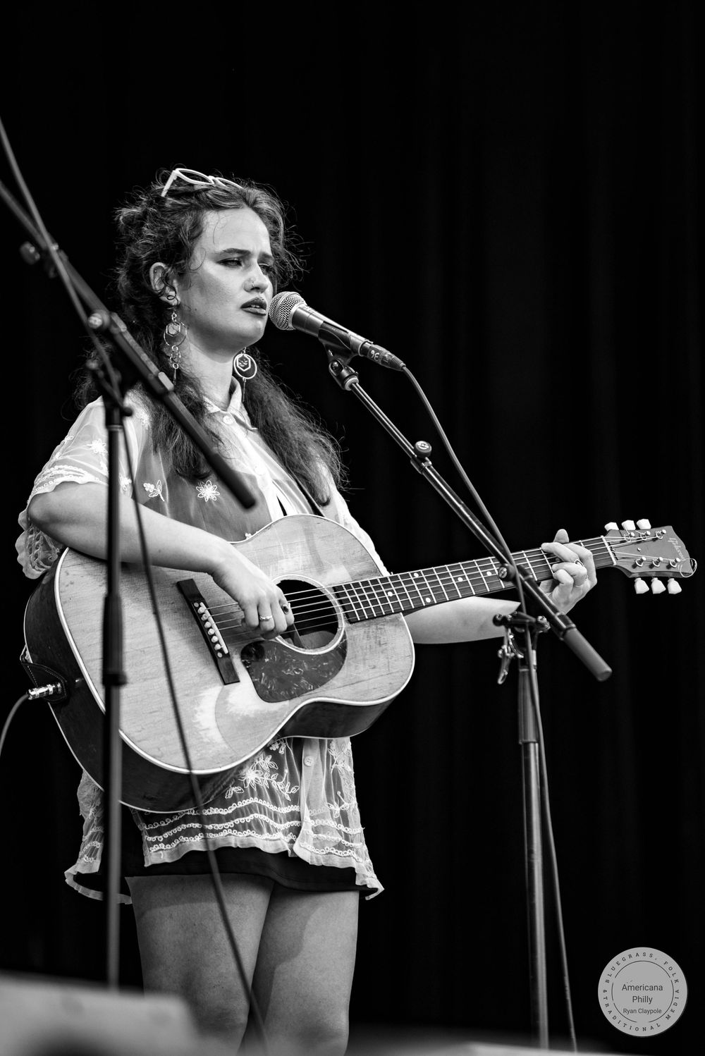 Young woman playing acoustic guitar and singing into microphone on stage, black and white concert photo.