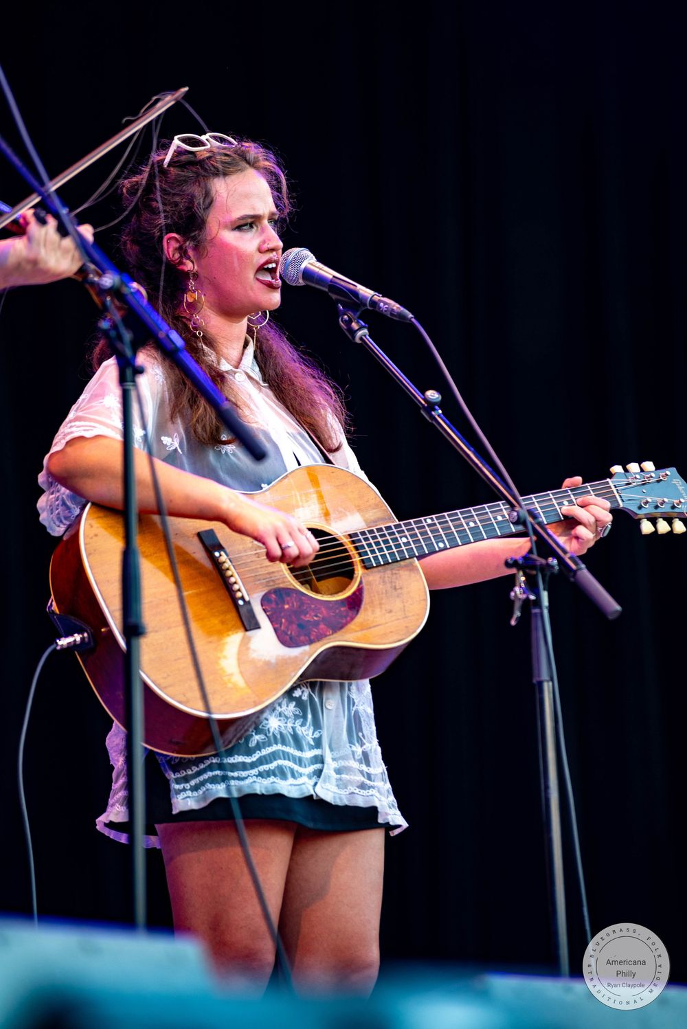 Young woman playing acoustic guitar and singing into a microphone on stage at an outdoor music festival.