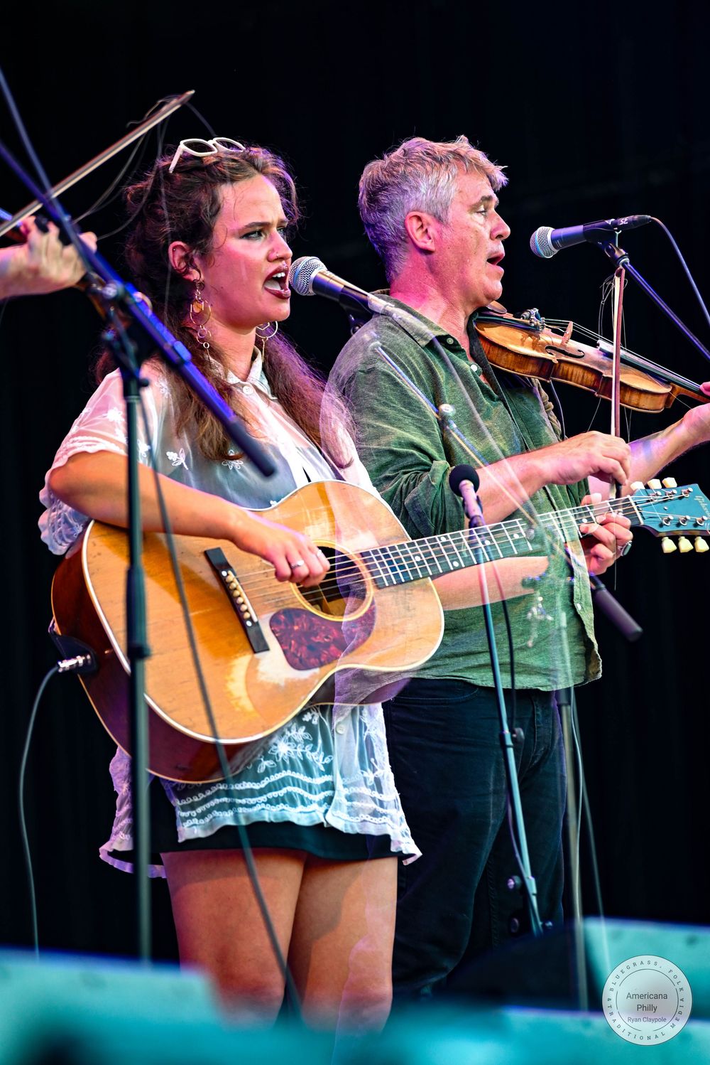 A woman plays acoustic guitar and sings while a man plays violin on stage at an outdoor music festival.