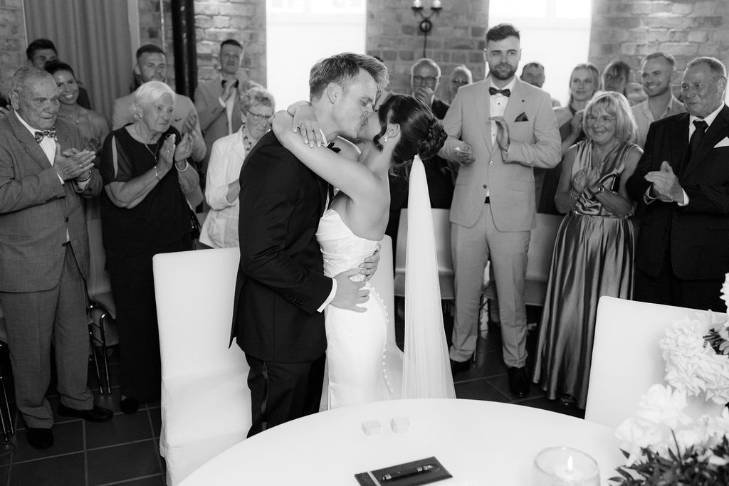 Black and white photo of a bride and groom sharing a first kiss at their wedding ceremony with guests applauding.