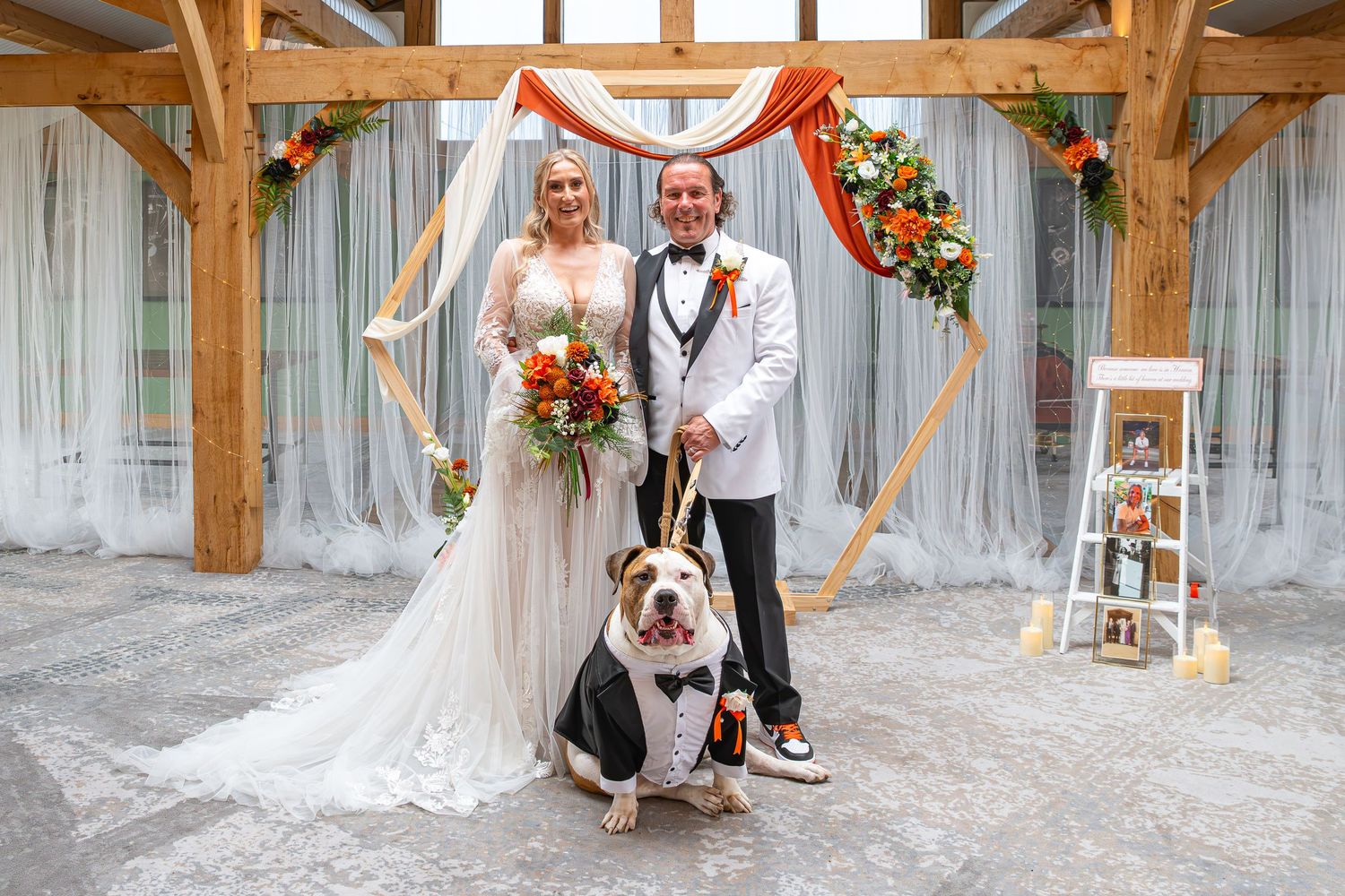 Bride and groom pose with their dressed bulldog under a hexagonal floral arch at an elegant fall wedding ceremony.