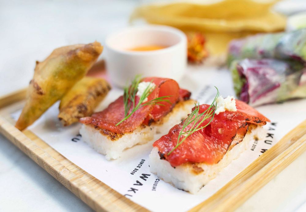 Elegant sushi dish topped with fresh tomato and herbs, served on a wooden board with dipping sauce.