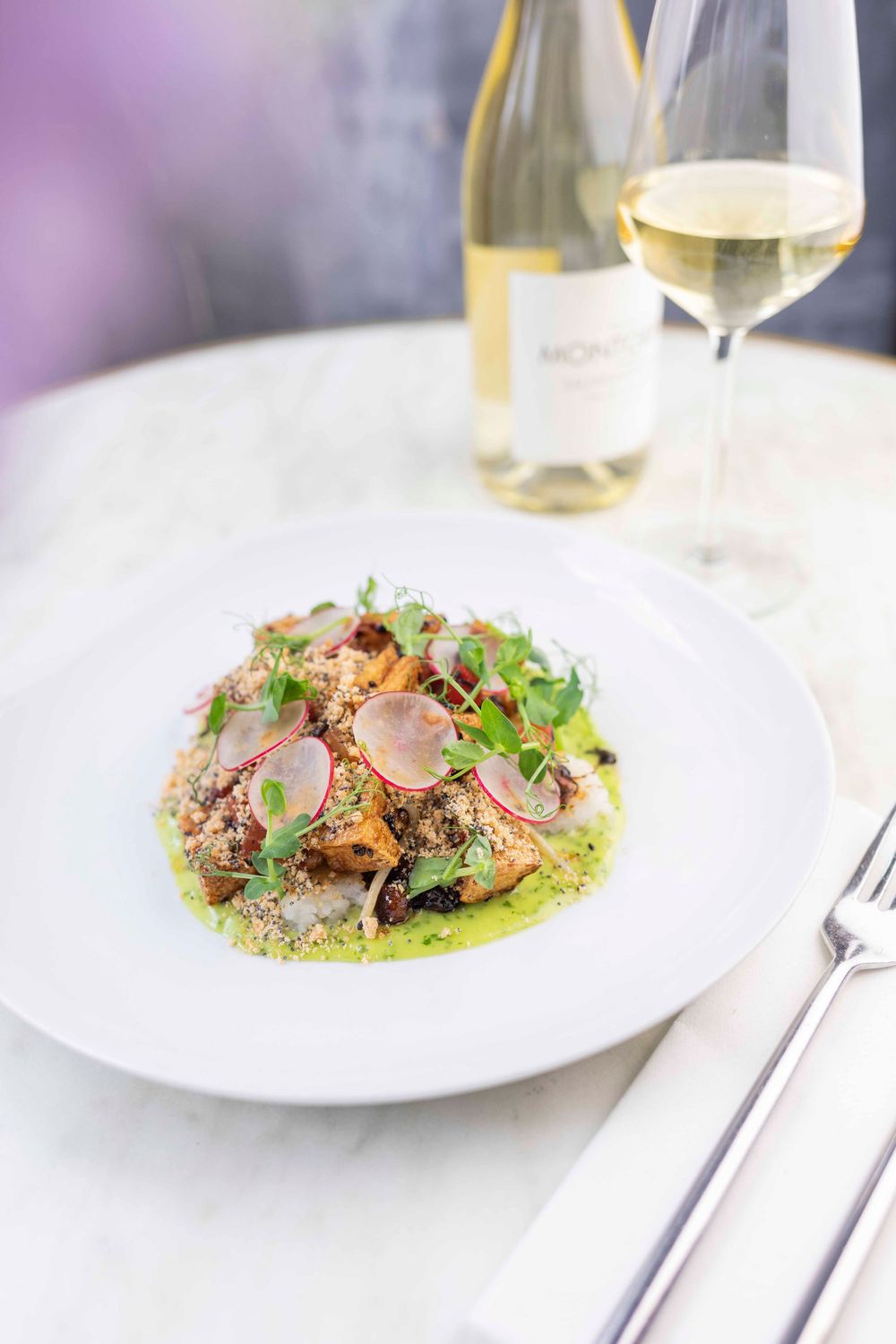 Elegant white plate with herb-crusted fish, radish garnish, green sauce, paired with white wine at a marble table.