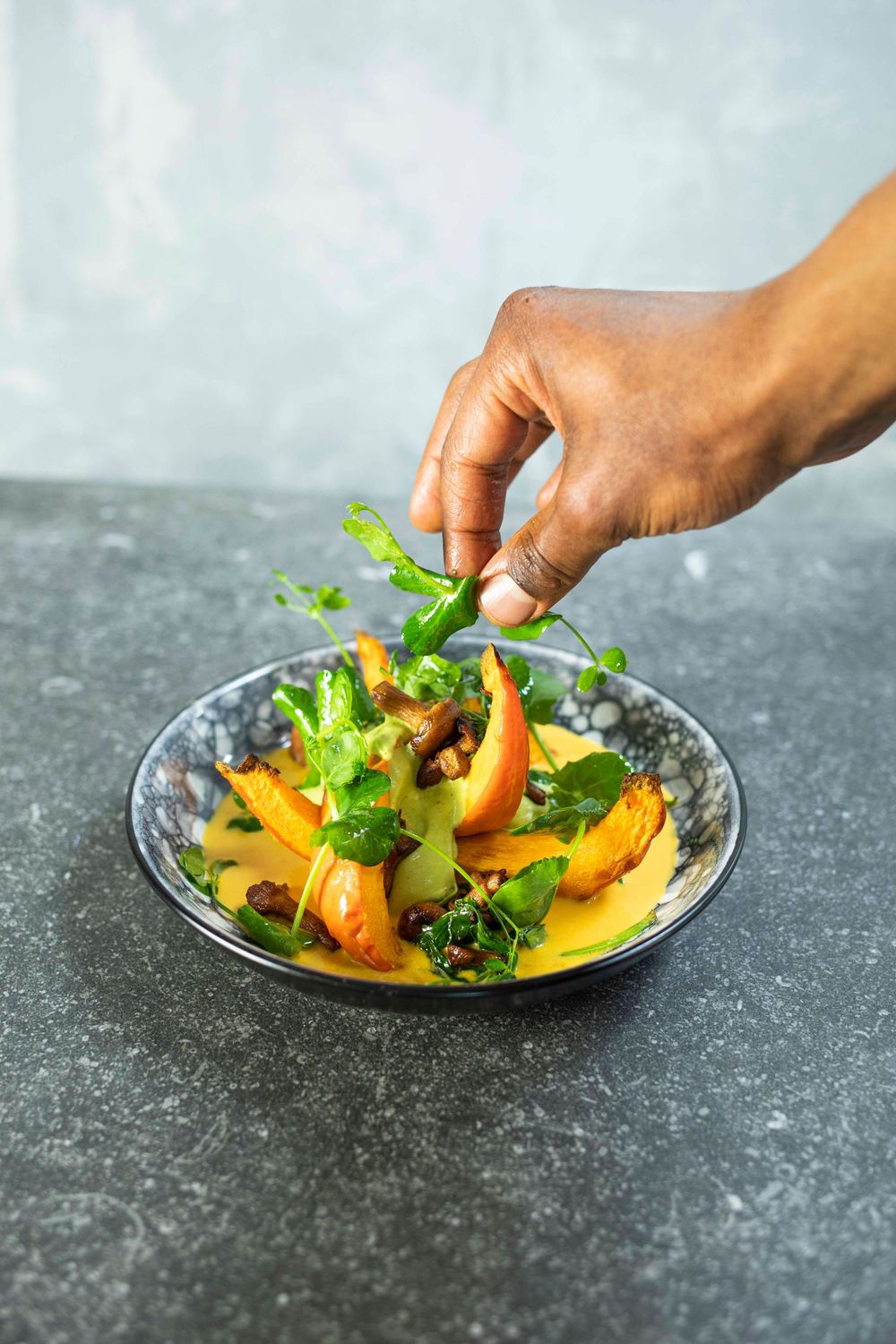 A hand garnishing a gourmet salad with greens, roasted vegetables, and mushrooms in a decorative bowl on a stone surface.