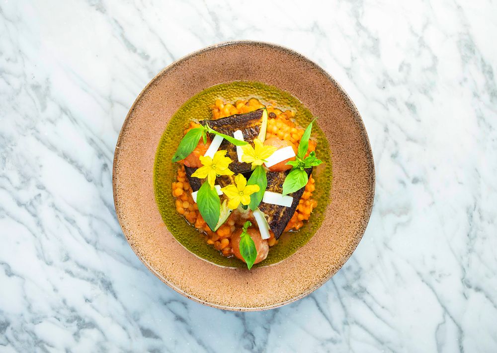 Gourmet dish with green herb sauce, chickpeas, fish, yellow flowers, and fresh basil on a rustic ceramic plate.