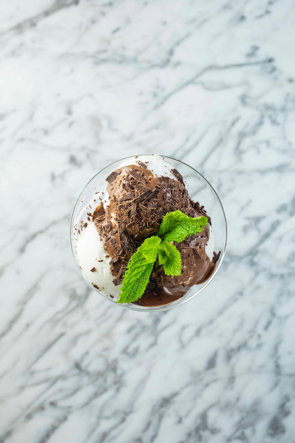 Glass dessert cup with vanilla and chocolate ice cream, chocolate shavings, and fresh mint leaves on marble surface.