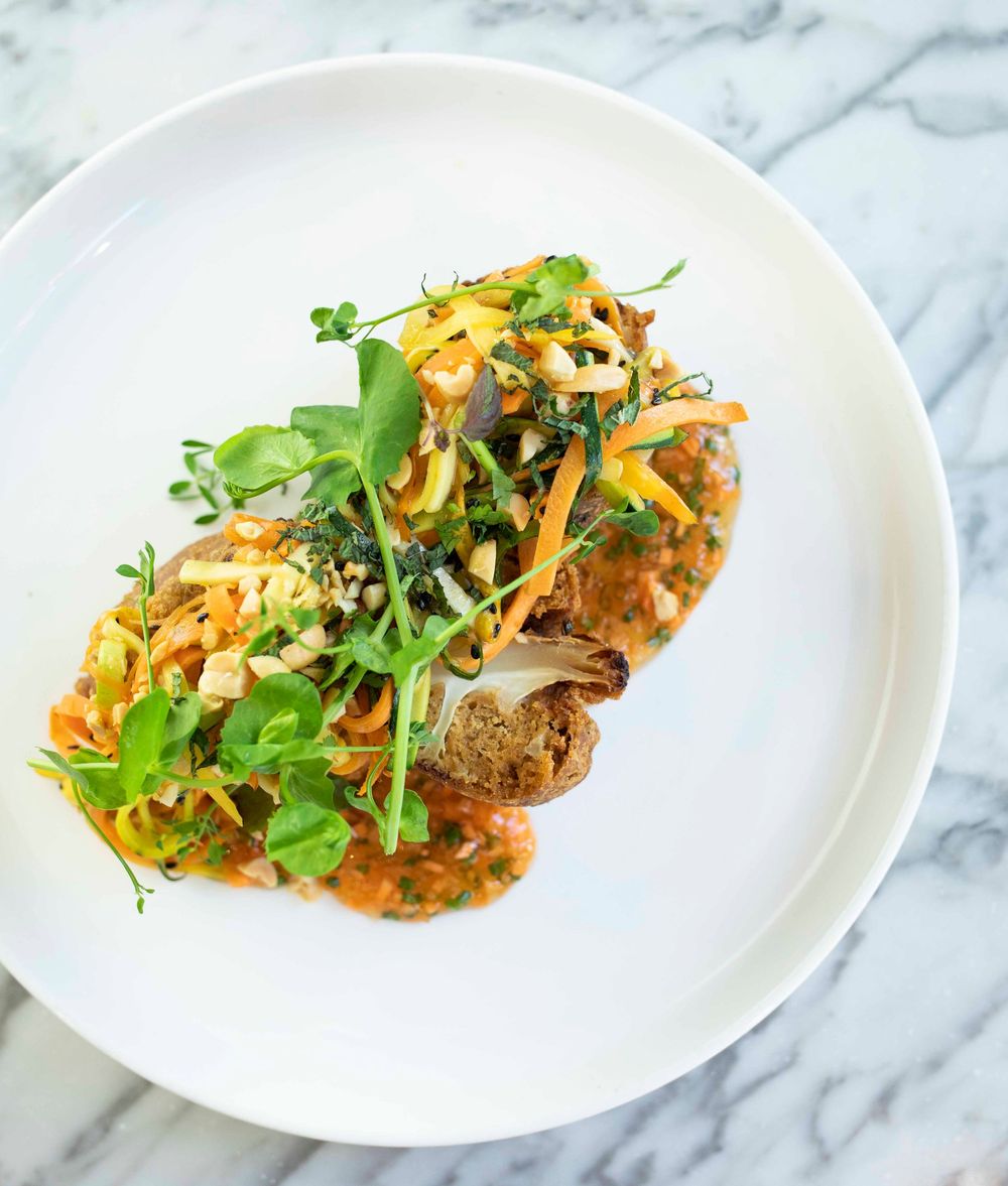 Gourmet open-faced sandwich topped with sautéed vegetables, microgreens, and tomato sauce on a white plate.