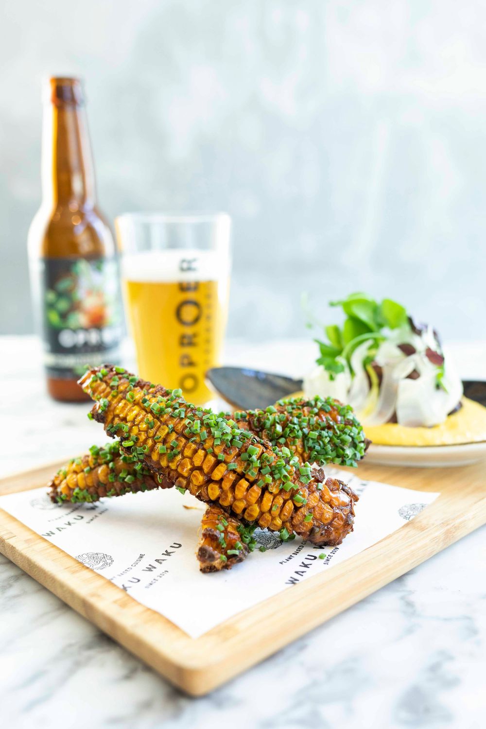 Grilled corn cobs topped with herbs on a wooden board, served with a cold beer and salad in the background.