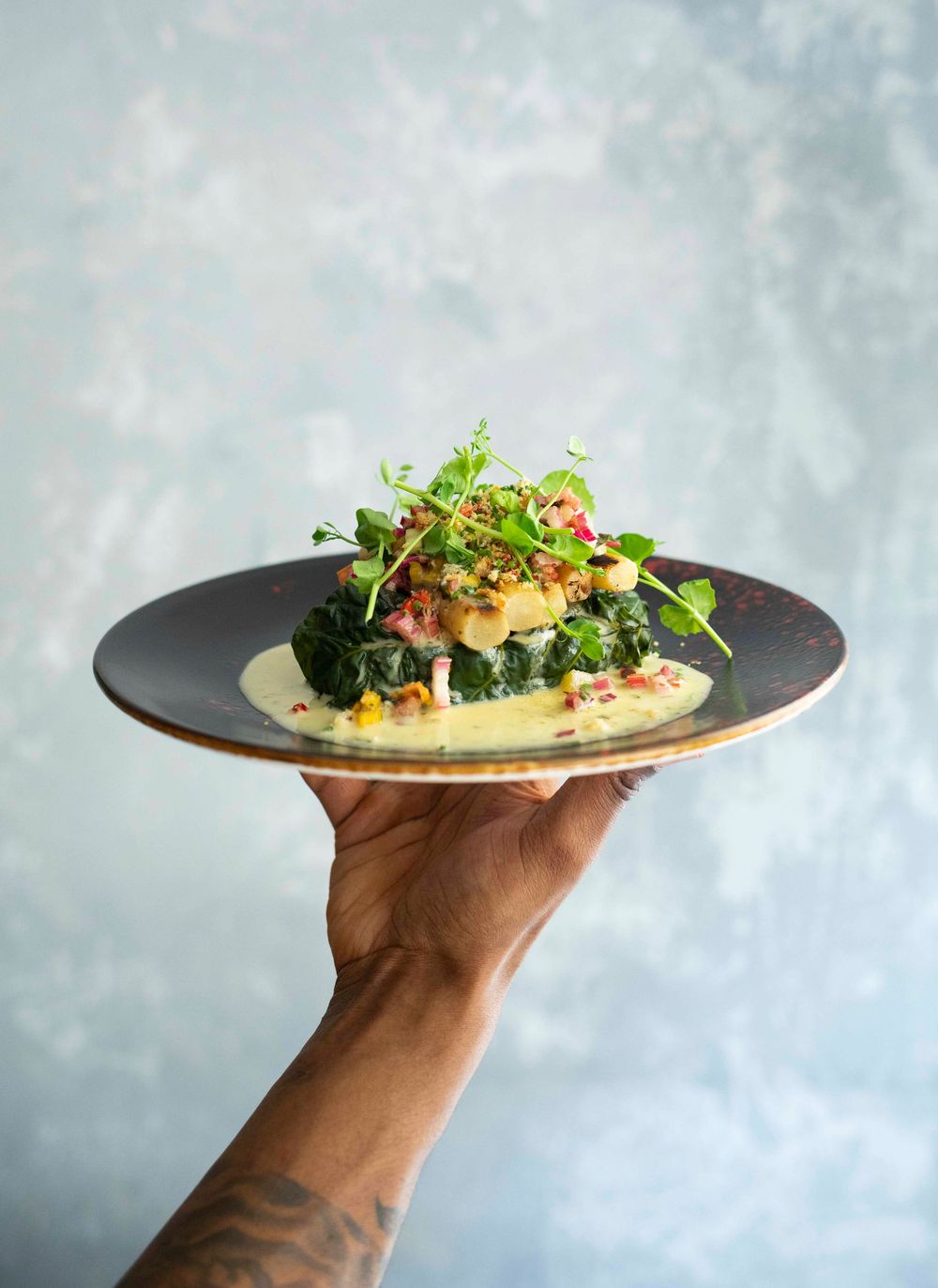 A tattooed hand holds a dark plate with an elegant stacked seafood dish topped with microgreens and cream sauce.