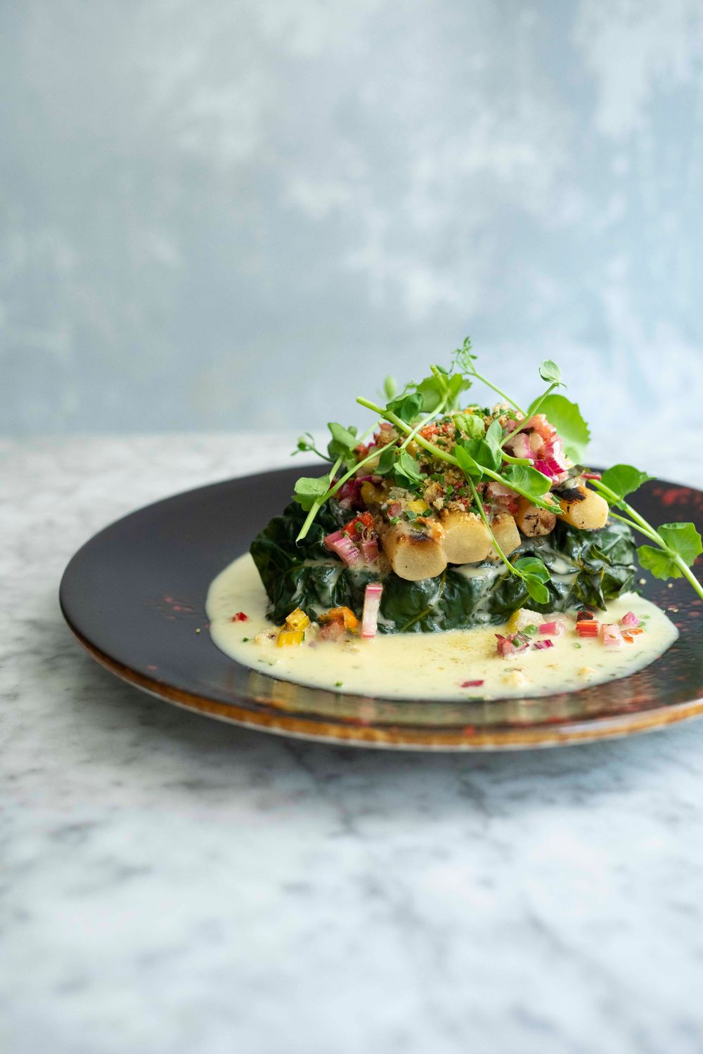 Elegant plated dish with sautéed greens, mushrooms, microgreens, and creamy sauce on a dark ceramic plate.