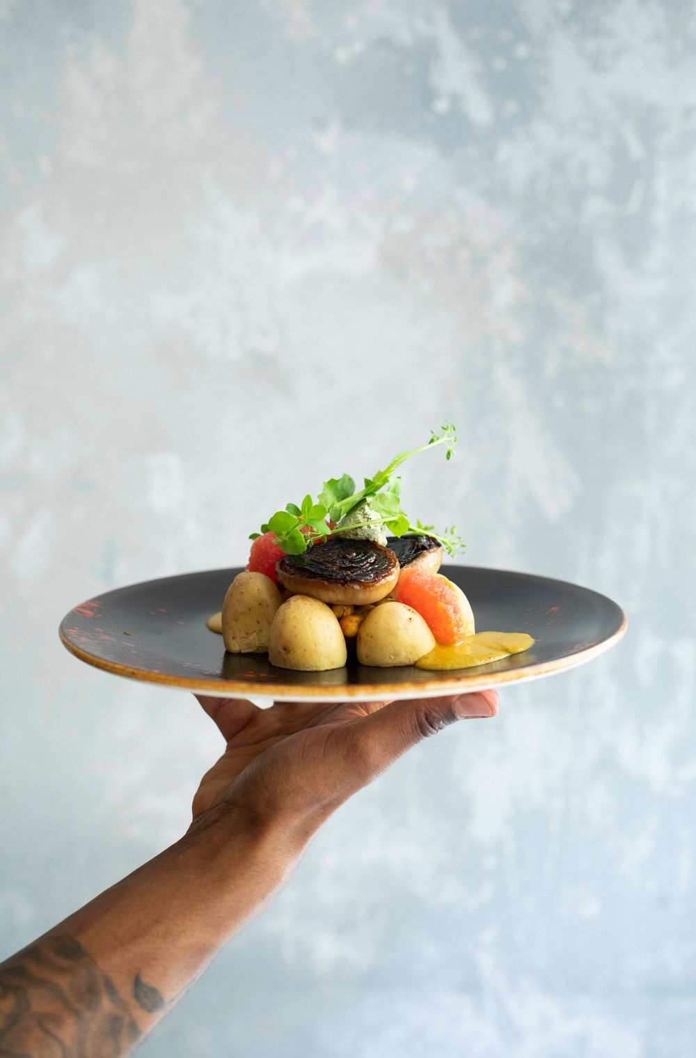 A tattooed hand holds a dark plate with gourmet dish of baby potatoes, tomatoes, mushroom, and fresh herbs.