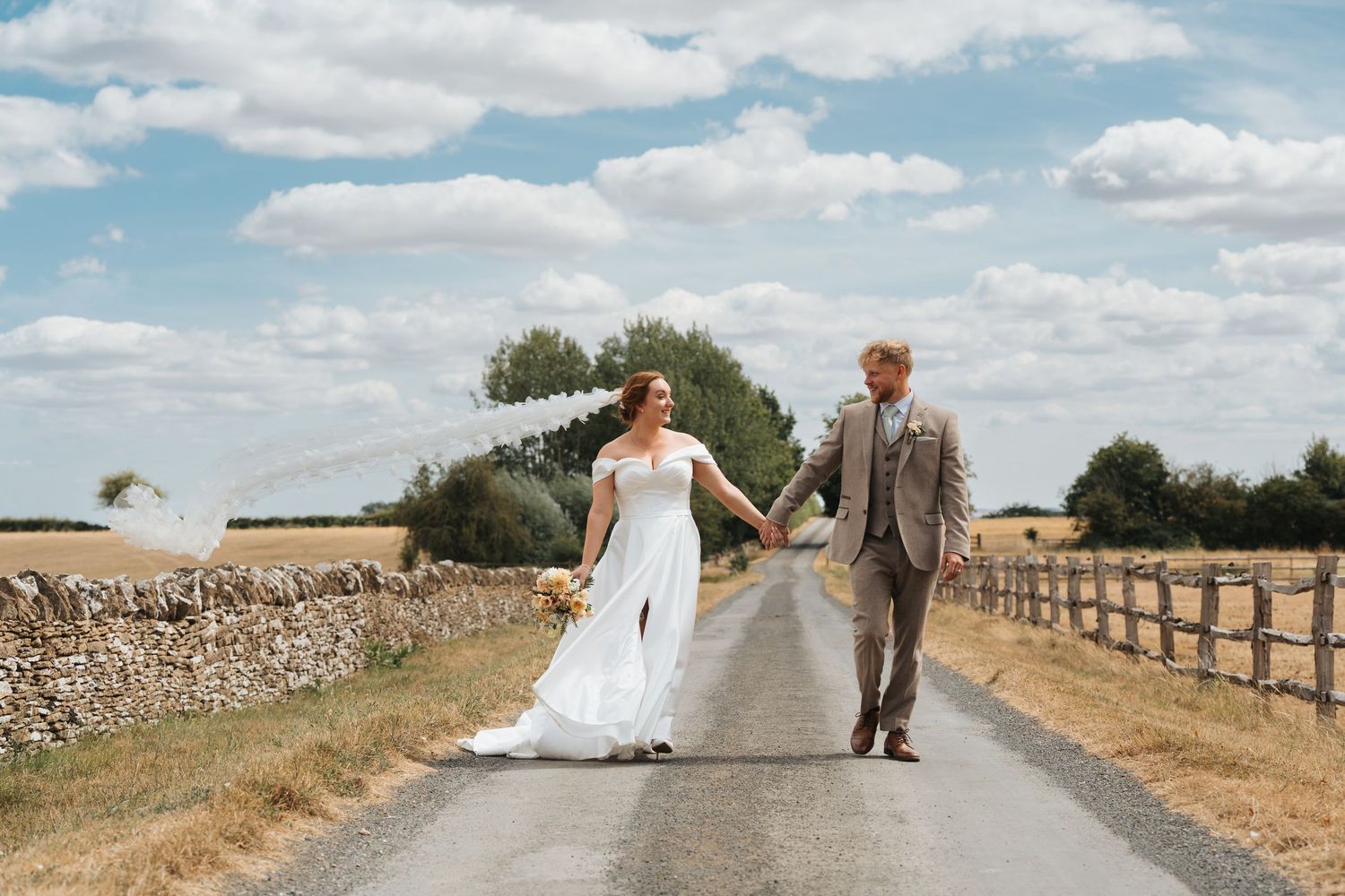 Bride and groom holding hands walking down a sunny country road on their wedding day.