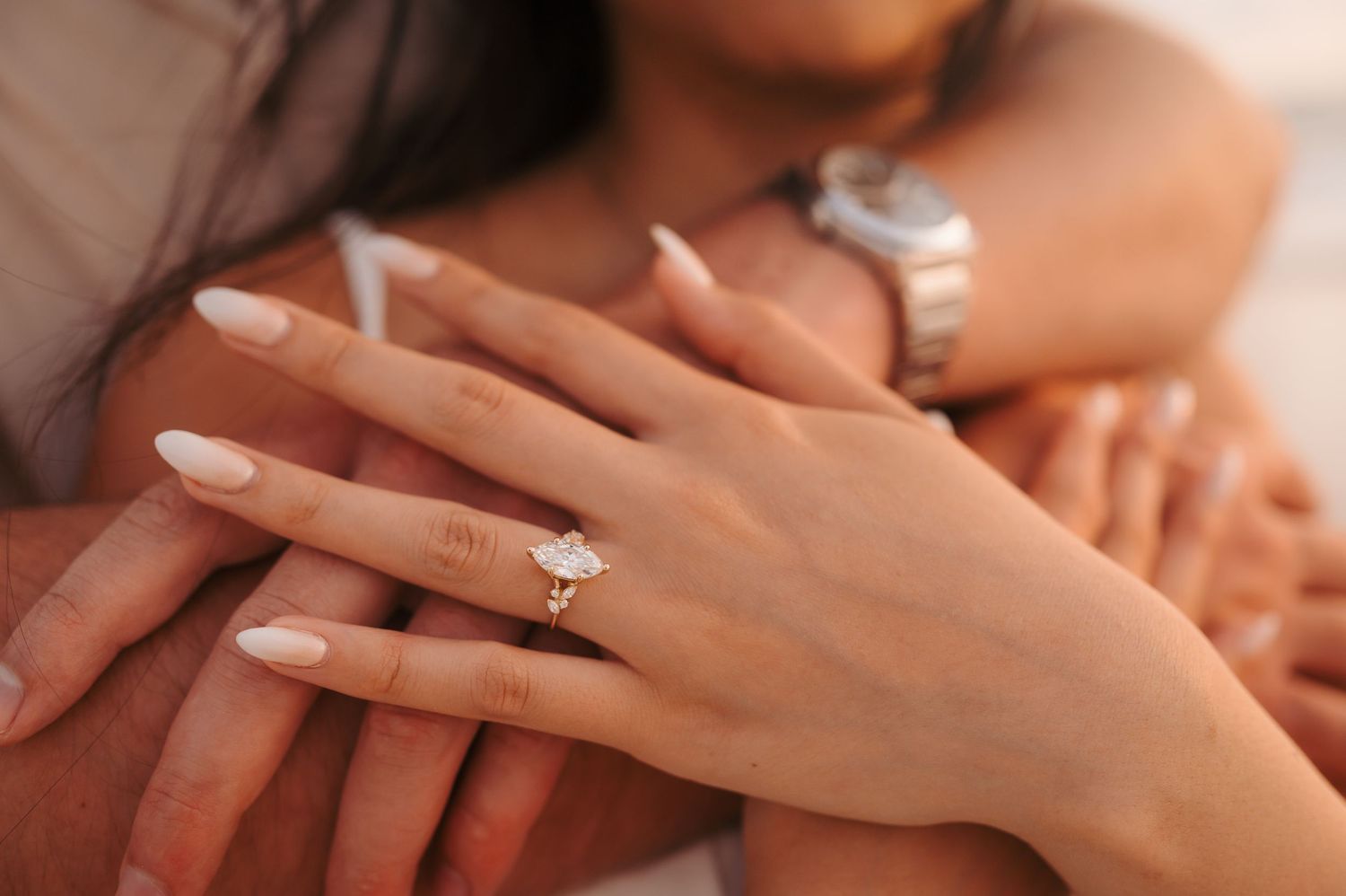 Close-up of a woman's hand wearing a diamond engagement ring resting on a man's arm with a watch.