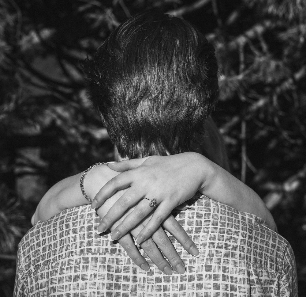 Black and white photo of a person embracing another from behind, showing a ringed hand on their shoulder.
