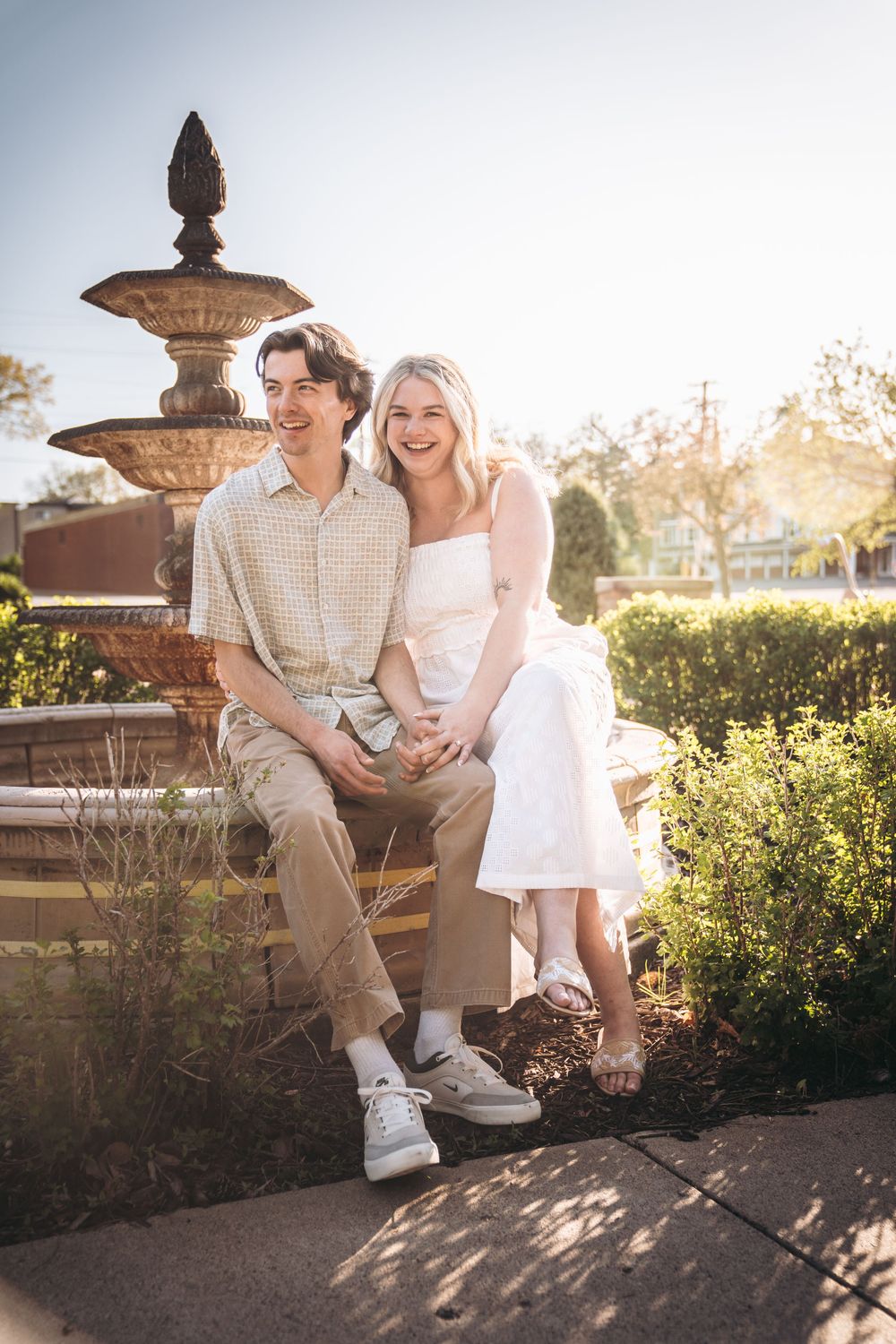 Happy couple sitting together by a vintage stone fountain in a sunny garden, man in plaid shirt and woman in white dress.