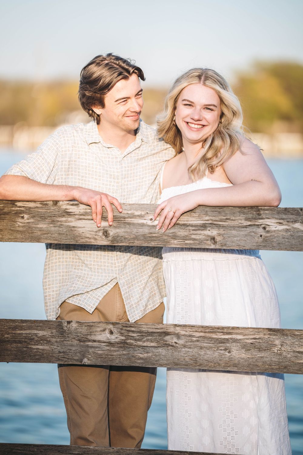 A smiling couple leans on a wooden dock railing by the water, the woman in white and man in a beige shirt.
