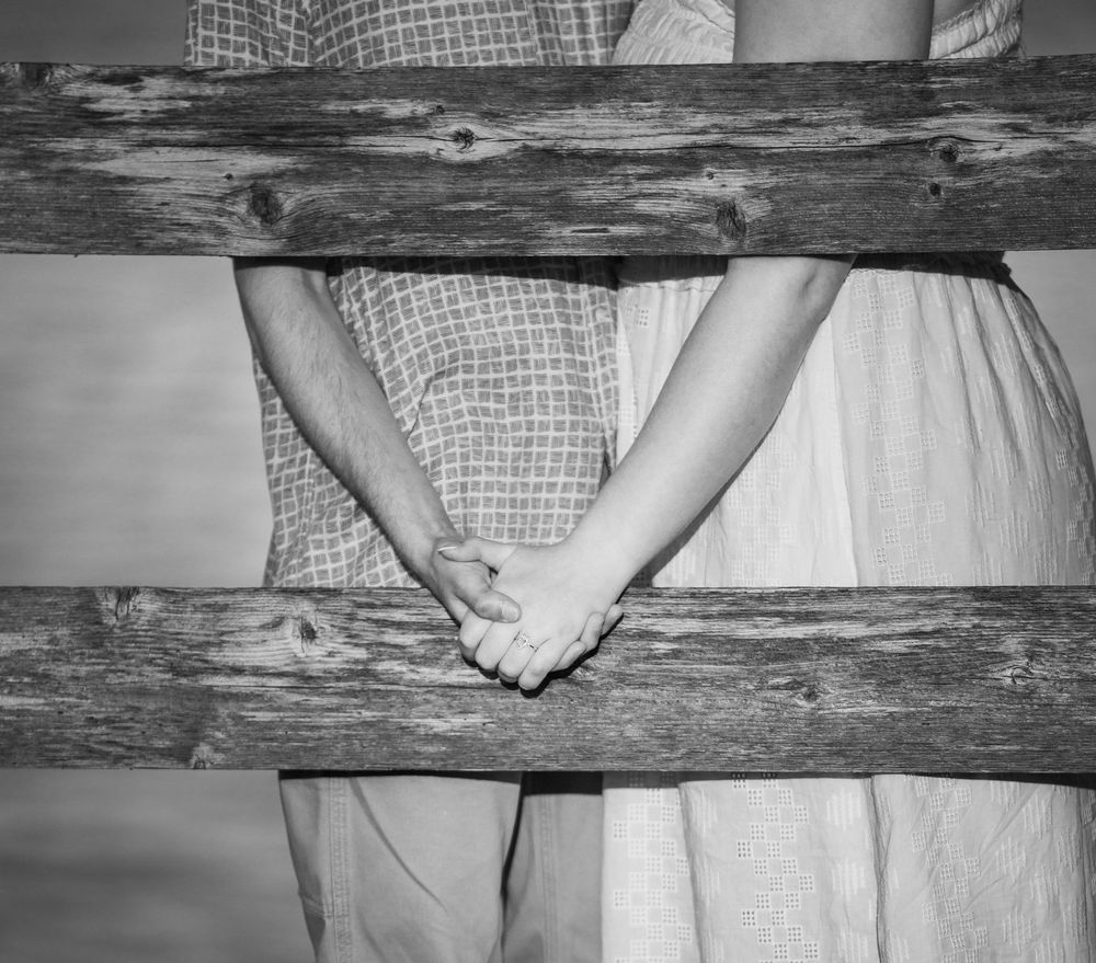 Black and white photo of a couple holding hands behind a rustic wooden fence, wearing casual country attire.