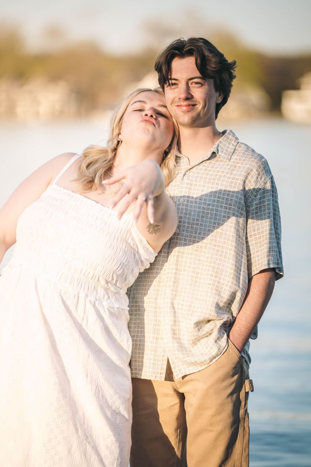Young couple standing by a sunlit lake, woman in white dress leaning on mans shoulder, smiling warmly together.