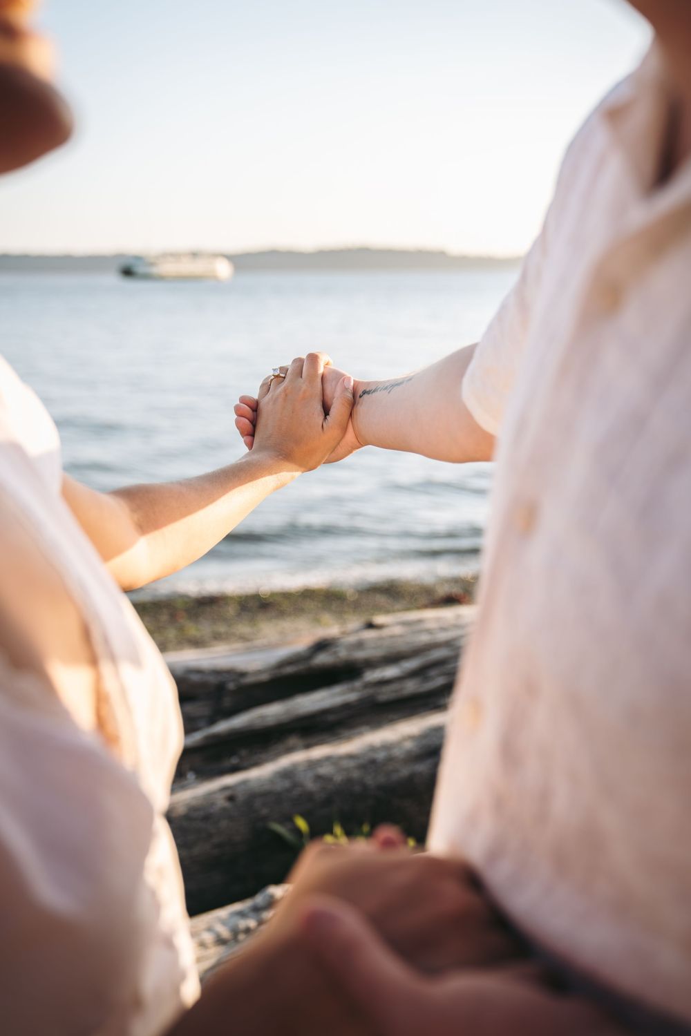 Two people hold hands by the water at sunset, wearing light-colored clothing near driftwood.